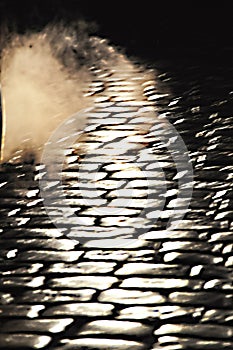 Cars drive on a cobblestone street at night, car lights on the street water vapor