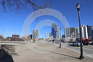 Cars driving along a street surrounded by glass office buildings, a wide smooth sidewalk, a tower crane and blue sky in downtown