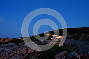 cars leaving the top of Cadillac Mountain after sunset with light streaks agains the alpen glow on pink granite boulders