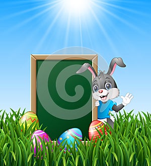 Cartoon bunny waving hand with easter eggs and green chalkboard in the grass