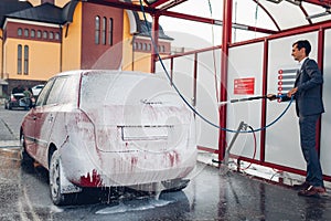 Carwash. Driver covers his auto with soap foam holding hose and cleans it with water.