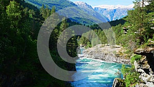 A cascading mountain stream rushes down through rock and forest with snow capped peaks and blue sky in the distance.