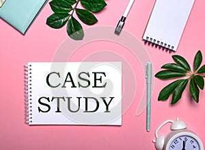 CASE STUDY is written in green on a white notepad on a pink background surrounded by notepads, pens, white alarm clock and green