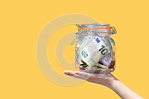 Cash euro banknotes in a glass jar in the hand of child isolated yellow background. Savings on education. Copy space