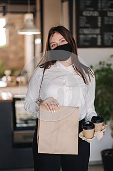 Cashier in face mask holding package with food and two cups of coffee. Contactless payments concept at cafe. Take away