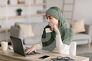 Cashless payment. Young Arab woman in hijab holding credit card and using laptop pc, shopping online from home