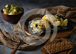 A casserole with baked fish and cheeze sauce on the dark background, potatoes in a clay bowl