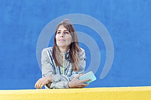 Casual adult woman lean on yellow wall holding a mobile phone while looking away outdoors against a blue background