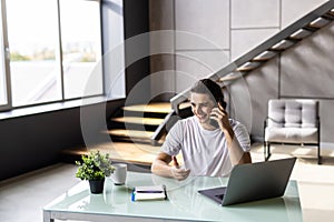 Casual businessman working at office desk, using mobile phone and laptop computer, typing, making phone call