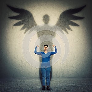 Casual confident woman flexing arm muscles as casting a hero shadow with angel wings on a dark room wall. Super power, inner