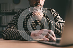 Casual contemplative adult male using laptop computer in dark home office interior