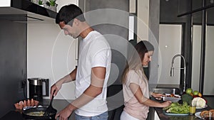 Casual couple making morning breakfast together in domestic kitchen. Positive pretty woman chopping salad and handsome