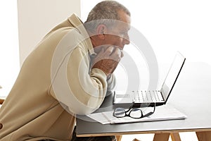 Casual dressed and anxious man biting his fingers as he looks at a computer screen. A pair of glasses lying on a pile of papers ne