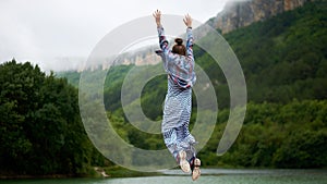 Casual dressed hiking woman under the rain jumping near the lake in mountains