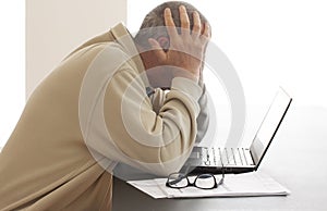 Casual dressed man is bending his head over computer in despair while he hides his head in his hands. Pair of glasses lying on a p