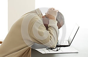 Casual dressed man bends his head over computer in despair while he hides his head in his hands. Pair of glasses lying on a pile o