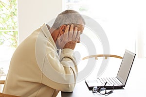 Casual dressed man sitting by a laptop computer is hiding his head in despair. A pair of glasses lying on a pile of papers next to