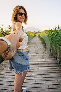 Casual dressed woman walking in nature at sunset, holding my hand in first person and smiling