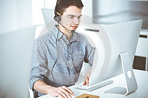 Casual dressed young man using headset and computer while talking with customers online. Call center, business concept