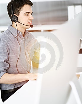 Casual dressed young man using headset and computer while talking with customers online in sunny office. Call center