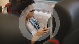 Casual female passenger on the train sits in a comfortable seat using smartphone e-commerce app, looking at the window.