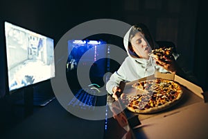 Casual gamer sits at a table at home near a computer and eats delicious pizza out of the box. Hungry young man relaxing sitting at