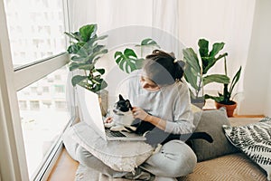 Casual girl working on laptop with her cat, sitting together in modern room with pillows and plants. Cute cat helping owner during