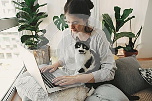 Casual girl working on laptop with her cat, sitting together in modern room with pillows and plants. Home office. Cute cat helping