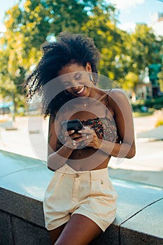 Casual hipster Afro-Columbian woman sitting outdoors on a small wall texting on her mobile phone