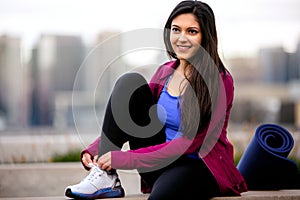 Casual lifestyle portrait of beautiful Indian American woman relaxing after cardio fitness exercise, city skyline buildings in bac