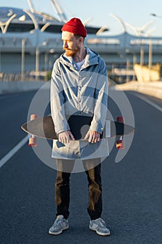 Casual man with longboard over city building. Hipster stylish skateboarder hold skateboard on sunset