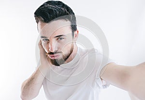 Casual man taking selfie in studio alone. Look straight and pose. Wear white shirt. Bearded man isolated over white