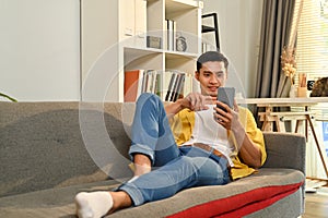 Casual millennial asian man relaxing on couch at home texting messaging, browsing wireless internet on smartphone