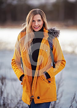 Casual portrait of young woman in warm orange yellow winter jacked, zipper down, smiling, blurred snow covered landscape behind