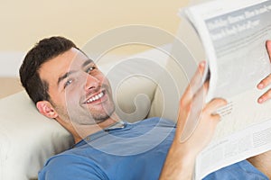 Casual smiling man lying on couch holding newspaper