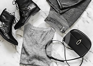 Casual women`s clothing - grey mom`s jeans, cashmere pullover, black boots, cross body bag on a marble background, top view.