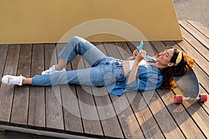 Casual young hipster girl in sunglasses texting in smartphone outdoors lying relaxed in longboard