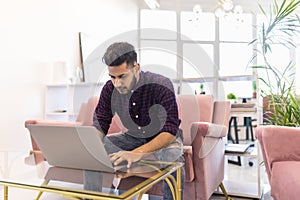 Casual young indian man using laptop with happy on sofa in modern office