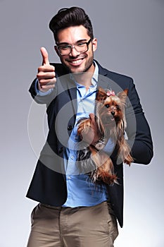 Casual young man holds puppy and shows thumb up