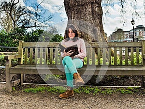 A Casual Young Woman Sitting on a Bench in a Park and Reading