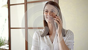 Casual young woman in white blouse standing in living room smiling laughing talking on phone