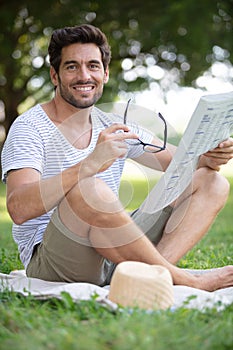 casually dressed man sat on grass reading newspaper