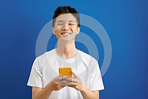 Casually dressed student standing isolated on blue background looking at screen of smart phone, browsing web pages and smiling