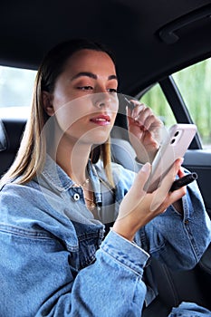 Casually Dressed Woman Putting On Make Up In Back Of Taxi Using Mobile Phone As Mirror