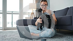 Casually dressed woman sits on a carpet with a laptop, holds on her knees and strokes a fluffy cat and works in a cozy