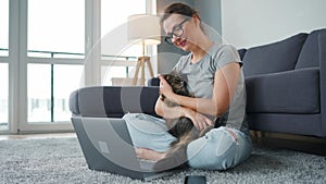 Casually dressed woman sits on a carpet with a laptop, holds on her knees and strokes a fluffy cat and works in a cozy
