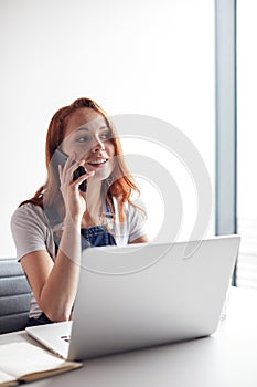 Casually Dressed Young Businesswoman Working On Laptop At Desk In Modern Workplace Making Phone Call