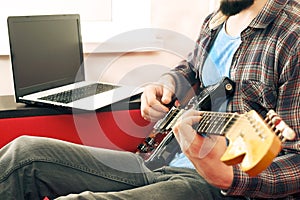 Casually dressed young man with guitar playing songs in the room at home. Online guitar lessons concept. Male guitarist practicing