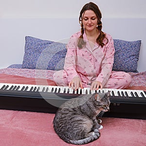 A cat sits next to a woman in red pajamas playing electric piano keys