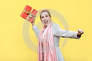 Catch your gift! Young adult crazy woman swung and wants to throw off your gift box, isolated on yellow background.